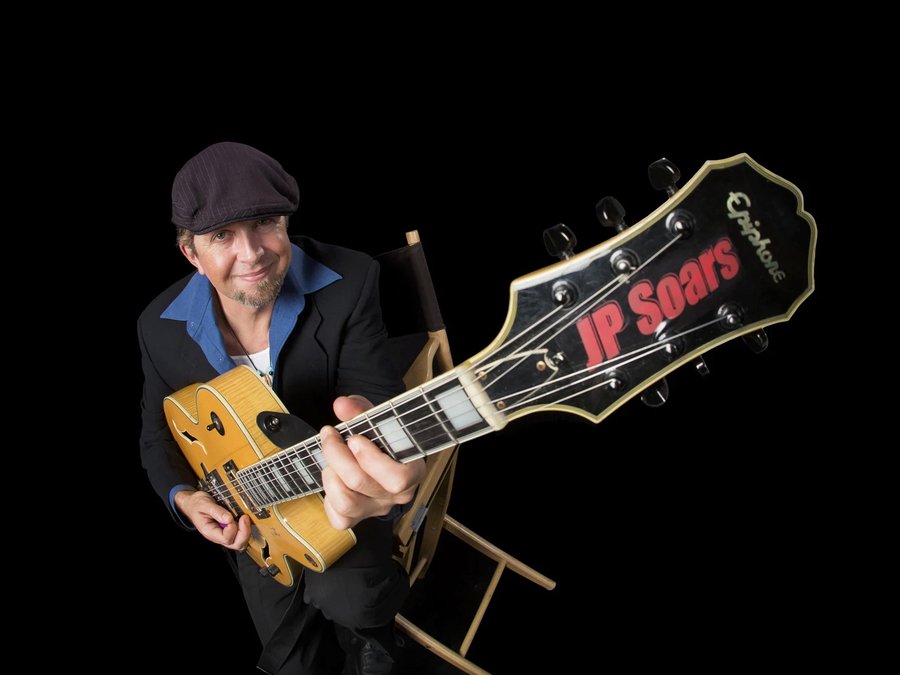 Guitarist holding a hollow-body guitar toward the camera against a black background